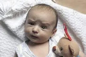 Head and shoulders of baby, lying on white blanket, holding a teddy. The baby is looking quizzically at the camera.